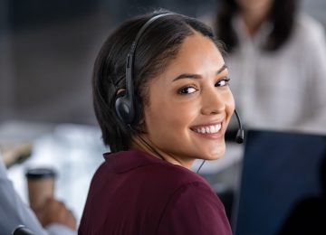 Retrato de una joven empresaria sonriente con auriculares en la oficina. Retrato de una agente de atención al cliente mirando a cámara. Feliz operador telefónico chica latina con auriculares trabajando en la computadora.